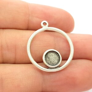 May include: A silver-coloured pendant setting featuring a circular frame and a smaller, round bezel. The bezel is designed to hold a small stone or cabochon. The pendant has a loop at the top for attaching to a chain or cord. The setting is held in a hand.