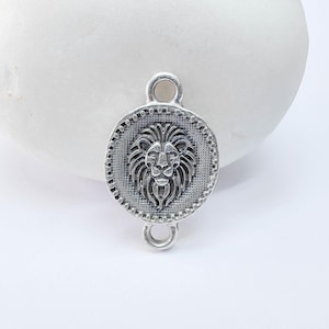 May include: A silver-toned metal charm with a lion's head design. The charm is circular with a beaded border and two loops for attachment. The lion's face is detailed with a mane and facial features.