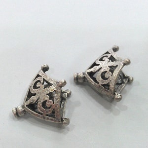 May include: Two silver-toned metal decorative pieces with an intricate, openwork design. Each piece is shaped like a square with rounded corners and has a delicate, floral pattern.