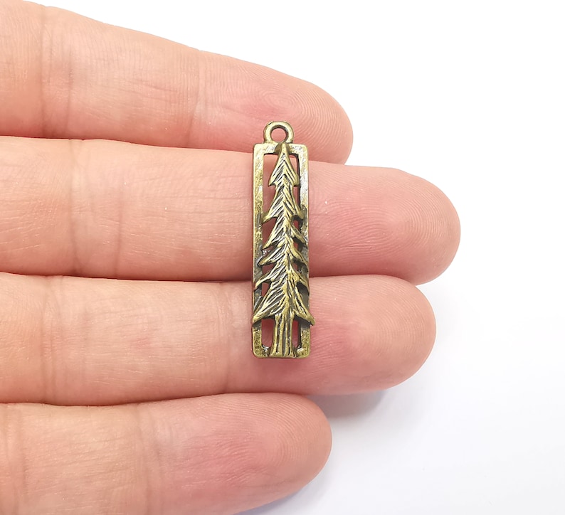 K&ouml;nnte beinhalten: Ein goldfarbener Metallanh&auml;nger in Form einer Tanne. Der Anh&auml;nger ist rechteckig und hat ein ausgeschnittenes Design einer Tanne. Der Anh&auml;nger hat oben eine Schlaufe zum Anbringen an einer Halskette oder einem Armband.