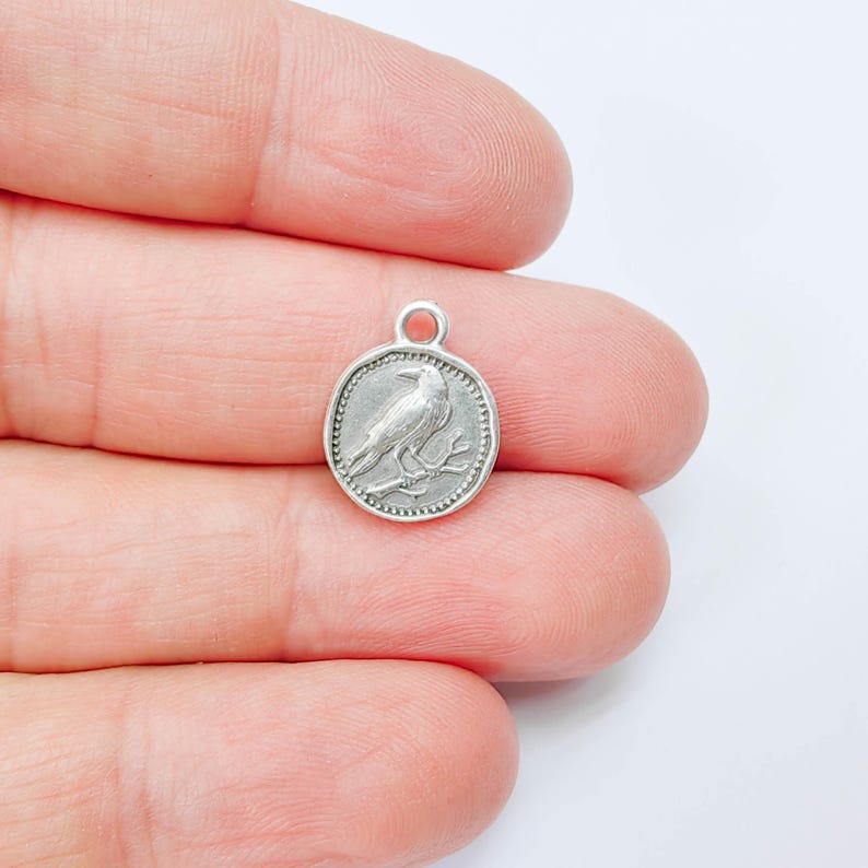 K&ouml;nnte beinhalten: Ein kleiner, runder, silberfarbener Anh&auml;nger mit einem detaillierten Relief eines Raben, der auf einem Ast sitzt. Der Anh&auml;nger hat oben eine Schlaufe zum Befestigen an einer Halskette oder einem Armband. Der Rabe blickt nach links, das Design ist von einem Perlenrand umgeben.