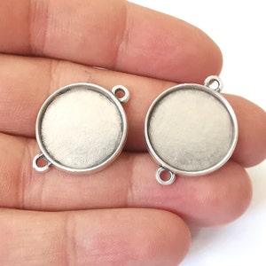 May include: Two silver-toned round metal bezel settings with a smooth, white surface. The bezels have two loops on the side for attaching to a chain or cord.