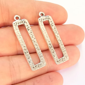 Puede incluir: Dos blanks rectangulares para pendientes de metal plateado con textura martillada. Los blanks tienen un agujero en la parte superior para sujetar los ganchos de los pendientes.