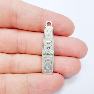 Puede incluir: Un colgante de metal plateado con forma rectangular alargada y esquinas redondeadas. La superficie está grabada con un diseño central de polilla, flanqueado por lunas crecientes arriba y abajo, y decorado con pequeños motivos de estrellas.
