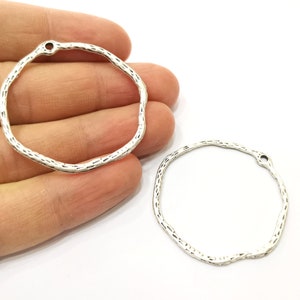 Op de afbeelding: Twee zilverkleurige metalen ringen met een getextureerd oppervlak. Elke ring heeft een klein gaatje bovenaan om een ​​touwtje of ketting aan te bevestigen.