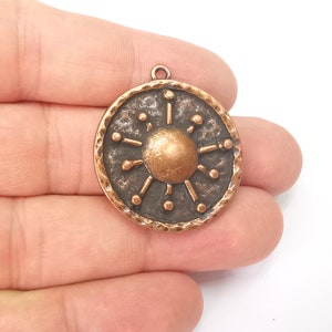 Op de afbeelding: Een koperkleurige hanger in de vorm van een schild met een zonnestralenontwerp. De hanger heeft een getextureerd oppervlak en een lus aan de bovenkant om een ​​ketting aan te bevestigen.