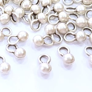 May include: A close-up of a pile of silver-toned metal bead caps with white pearl-like balls. The bead caps have a loop on each side.