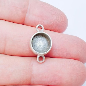 May include: A small, silver-coloured bezel pendant with a round setting. The setting is filled with a textured, silvery material. The pendant has loops at the top and bottom for attaching to a chain or other jewellery components.