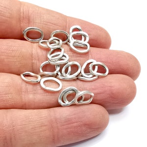 Może przedstawiać: Stosik srebrnych kółek do łączenia, o owalnym kształcie, z fakturą młotkową. Kółka mają średnicę około 1,2 cm.
