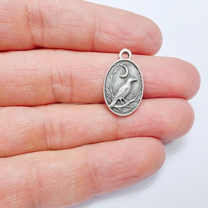 May include: Silver charm featuring a detailed design of a crow perched on a branch with a crescent moon in the background.