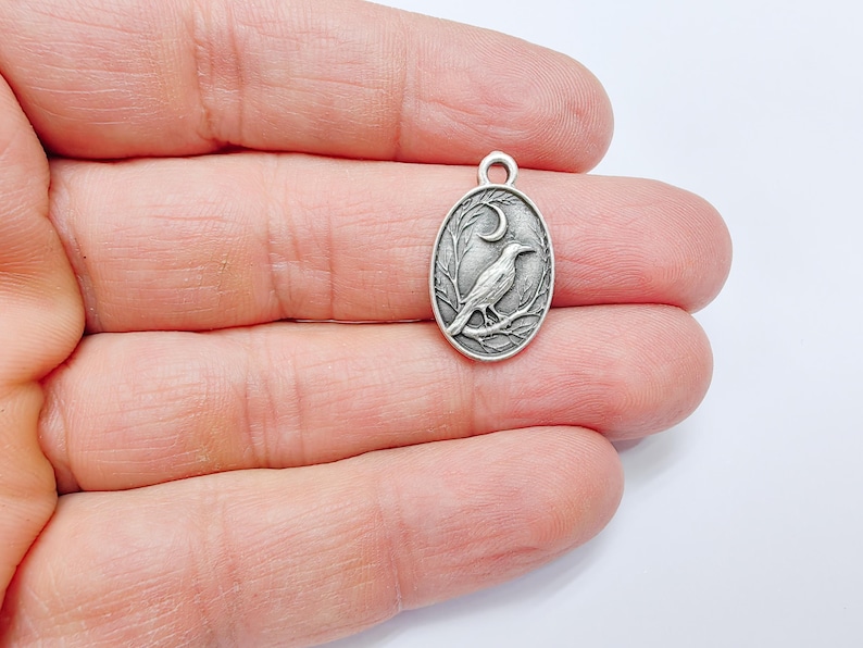 May include: Silver charm featuring a detailed design of a crow perched on a branch with a crescent moon in the background.