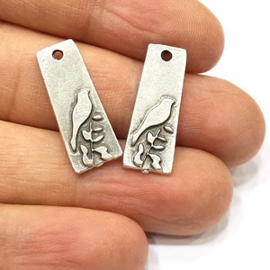 May include: Two silver-toned metal charms with a bird perched on a branch design. The charms are rectangular and have a hole at the top for attaching to jewelry.