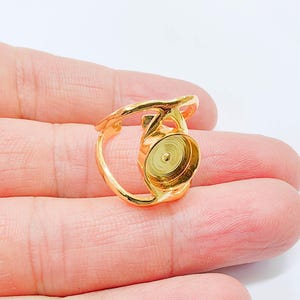 May include: A gold-coloured ring with a distinctive design. The ring has a circular setting with a textured interior, and an open, decorative band. The ring is held in a hand against a white background.