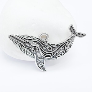 Op de afbeelding: Zilveren walvisbedel met ingewikkelde, draaiende ontwerpen. De walvis kijkt naar rechts en heeft een lus aan de bovenkant om aan een ketting of armband te bevestigen.