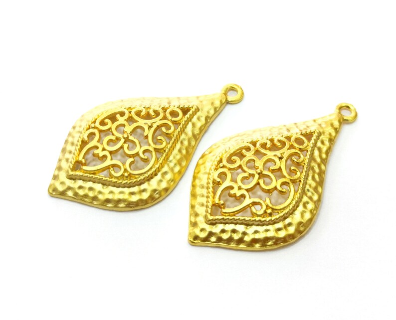 May include: Two gold-tone metal pendant charms with a filigree design. The charms are shaped like teardrops and have a textured surface.