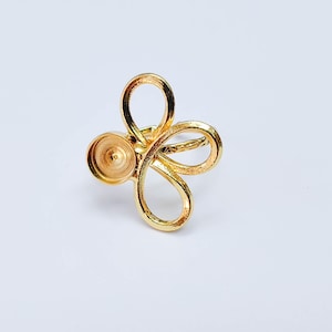 Op de afbeelding: Een goudkleurige metalen ringbasis met een open, lussenontwerp dat doet denken aan een gestileerde bloem of vlinder. De zetting bevat een cirkelvormig, verzonken kommetje bedoeld voor een cabochon of steen, bevestigd aan een gladde band.