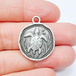 May include: A silver-coloured round pendant with a detailed sea turtle design. The turtle is surrounded by aquatic plants and bubbles, with a beaded border. The pendant has a loop at the top for attaching to a chain or bracelet.