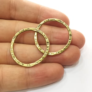Könnte beinhalten: Zwei strukturierte, messingfarbene Metallkreisanhänger. Die Anhänger haben ein gehämmertes Aussehen und werden vor einem weißen Hintergrund präsentiert. Dies sind wahrscheinlich Komponenten für die Schmuckherstellung oder das Basteln.