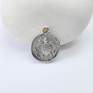 Può includere: Un ciondolo rotondo color argento con un design dettagliato di una tartaruga. Il ciondolo ha un bordo strutturato e un piccolo anello nella parte superiore per una catena. La tartaruga è circondata da piante acquatiche.