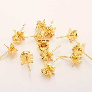 May include: A group of ten gold-toned earring posts with a decorative design. The posts are scattered on a white surface.