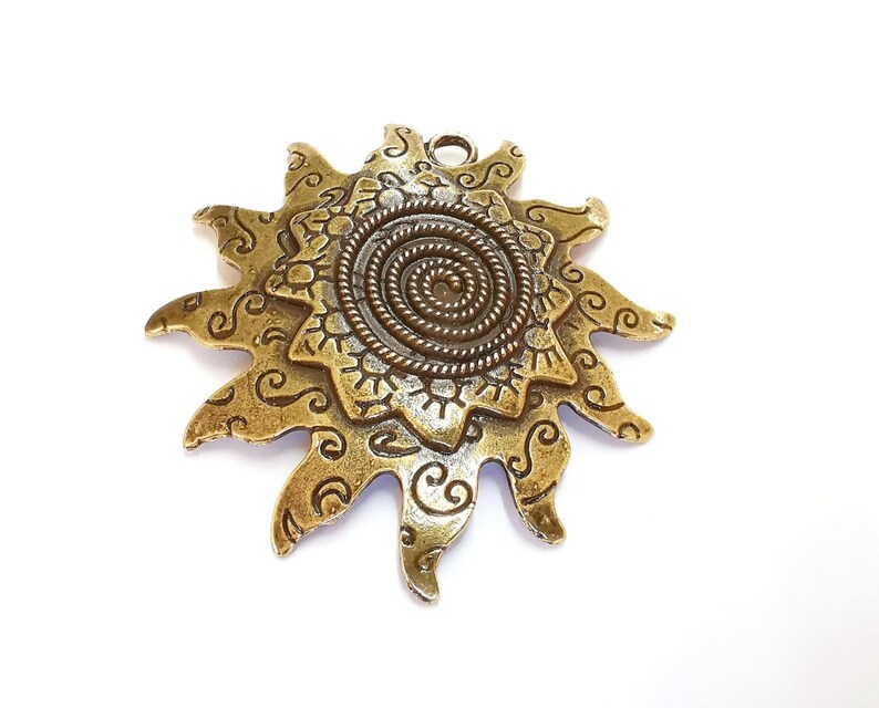 K&ouml;nnte beinhalten: Ein goldfarbener Metall-Sonnenanh&auml;nger mit einem detaillierten Spiraldesign in der Mitte und filigranen Ranken an den R&auml;ndern.