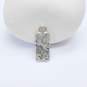Può includere: Un ciondolo rettangolare in metallo color argento con un design a libellula in rilievo su uno sfondo di canne e fogliame testurizzati. Il ciondolo include un occhiello circolare sulla parte superiore per l'aggancio, con una finitura dettagliata.