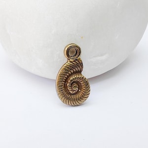 May include: A small, antique gold-colored pendant in the shape of a nautilus shell. The pendant has a spiral design with detailed ridges and a small loop at the top for a chain. The background is a white, smooth stone.