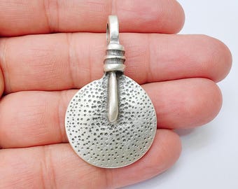 May include: A silver-toned pendant with a textured, circular design and a loop at the top for hanging. The pendant has a hammered appearance with small, evenly spaced indentations across its surface. The design includes a central, elongated element.