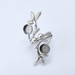 May include: A silver-toned ring with a botanical design. The ring features a spiraling band with leaf and berry accents, and two circular settings. The ring is made of metal and has a matte finish.
