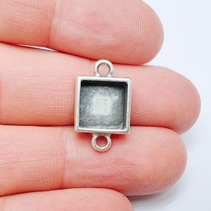 May include: A small, silver-coloured, square pendant with a recessed centre. The pendant has a loop at the top and bottom for attaching to a chain or other jewellery components. The pendant is held between fingers.