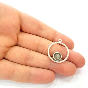 May include: A silver pendant featuring a circular frame with a smaller, textured circle within. It has a small loop at the top for attaching to a chain or other jewellery component. The design is simple and modern.