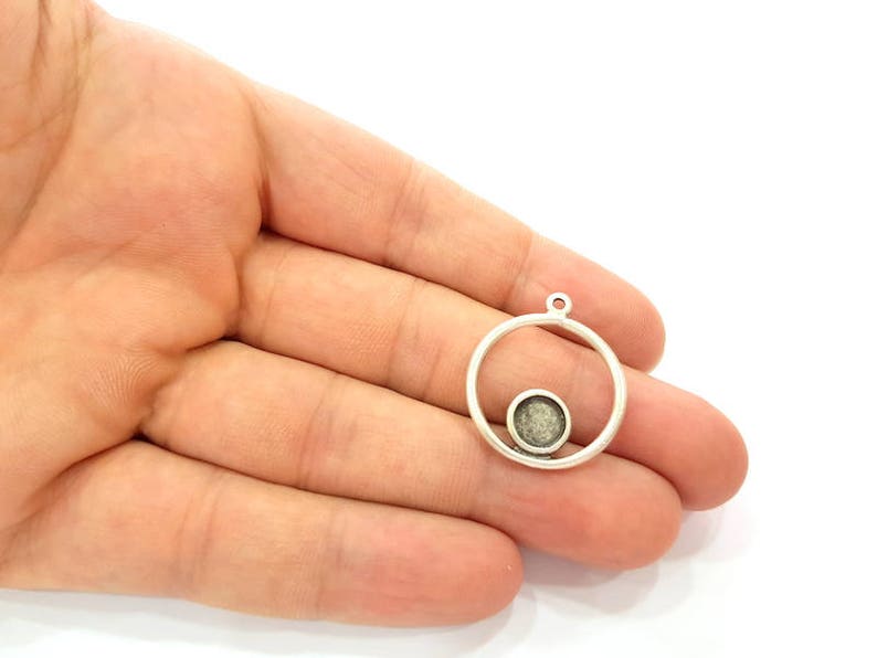 May include: A silver pendant featuring a circular frame with a smaller, textured circle within. It has a small loop at the top for attaching to a chain or other jewellery component. The design is simple and modern.