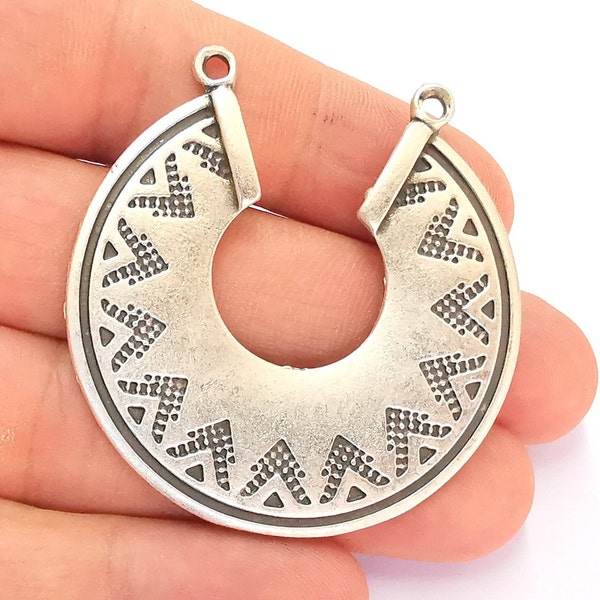 May include: Silver-toned metal pendant with a geometric design. The pendant is shaped like a half-circle with a hole in the centre. The design features a repeating pattern of triangles and dots.