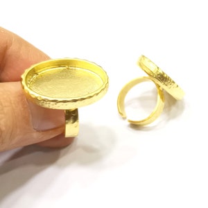 Op de afbeelding: Twee goudkleurige verstelbare ringen. De ringen hebben een ovale bezel-instelling met een getextureerde rand. Eén ring wordt vastgehouden. De andere ring ligt op een wit oppervlak.