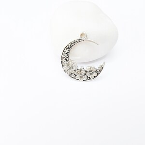 May include: A silver-toned crescent moon pendant with floral detailing. The moon shape is adorned with embossed flowers and intricate patterns. The pendant has a small loop at the top for hanging and is set against a white background.