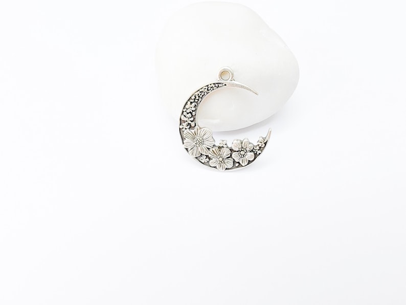May include: A silver-toned crescent moon pendant with floral detailing. The moon shape is adorned with embossed flowers and intricate patterns. The pendant has a small loop at the top for hanging and is set against a white background.
