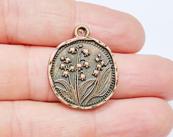 May include: A round, copper-colored charm with a detailed lily of the valley design. The charm features a small loop at the top for attaching to a necklace or bracelet. The edges are textured with a beaded border.
