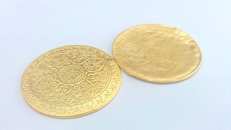 Peut inclure: Deux pendentifs ronds en m&eacute;tal de couleur dor&eacute;e. Un pendentif a un motif en relief d&eacute;taill&eacute; et l'autre est lisse.