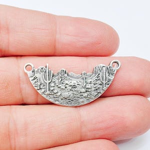 May include: Silver charm with a detailed design of a desert landscape with cacti and mountains. The charm has two jump rings for attaching to a necklace or bracelet.