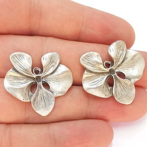 Może przedstawiać: Dwa srebrne charms w kształcie kwiatu orchidei o szczegółowym wzorze. Charms mają otwór u góry do przymocowania do biżuterii.