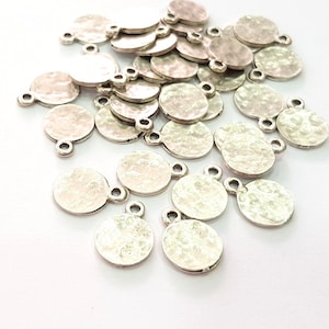 Op de afbeelding: Een stapel zilverkleurige ronde metalen bedels met een gehamerde textuur. Elke bedel heeft twee kleine lussen om aan sieraden of andere knutselwerkjes te bevestigen.