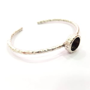 Puede incluir: Un brazalete de plata con textura martillada. El brazalete abierto presenta un engaste ovalado con interior oscuro. El metal brillante contrasta con el engaste.