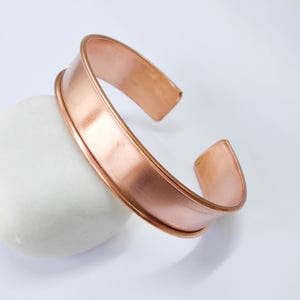 Peut inclure: Un bracelet manchette en cuivre au design simple et élégant. Le bracelet a une surface lisse et polie et une forme légèrement incurvée. Les extrémités ouvertes permettent de le porter facilement. Le bracelet repose sur une pierre blanche.