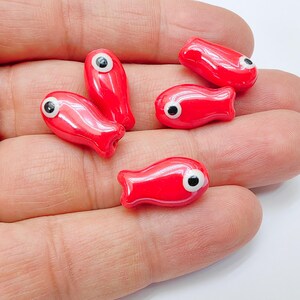 Pode incluir: Cinco contas de vidro vermelho em forma de peixe com olhos brancos e pretos. As contas têm a forma de um peixe e um acabamento brilhante.