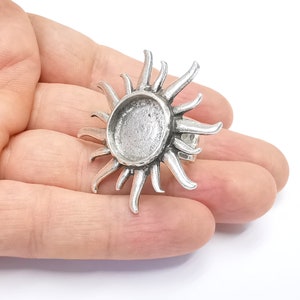 May include: A silver-colored ring with a sunburst design. The ring features a central, circular setting, surrounded by pointed rays. The ring is held in a hand against a white background.