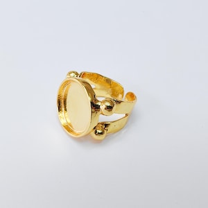 May include: A gold-coloured adjustable ring with an oval bezel setting. The ring features a double-band design with decorative ball accents. The setting is designed to hold a cabochon or other decorative element.