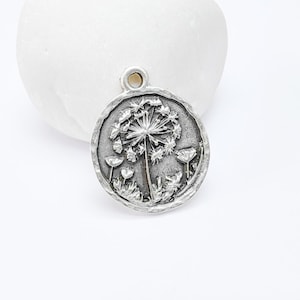 Puede incluir: Encanto de plata con un diseño en relieve detallado de una flor de diente de león con semillas que se dispersan en el viento.