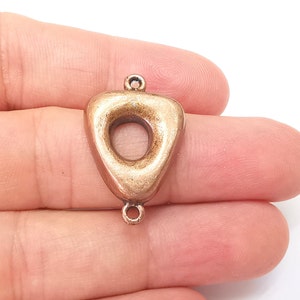 Puede incluir: Un amuleto de metal de color dorado en forma de triángulo con un círculo grande abierto en el centro. El amuleto tiene dos bucles en la parte superior para sujetarlo a joyas.