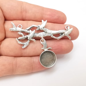 May include: A silver-toned metal charm featuring a branch with three birds and a round bezel setting. The charm has a rustic, textured finish and small loops for attaching to jewelry or other crafts.