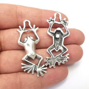 Pode incluir: Dois pingentes de sapo de prata com detalhes elaborados. Os sapos est&atilde;o em posi&ccedil;&atilde;o elevada com as pernas esticadas. Os pingentes t&ecirc;m um la&ccedil;o na parte superior para prender a joias ou outros artesanatos.